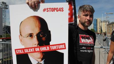 A War on Want campaigner at a demonstration holds a placard that has an image of G4S CEO Ashley Almanza's face with the words "Still silent on child torture" over his mouth, and #StopG4S in the top-right corner. Credit: War on Want