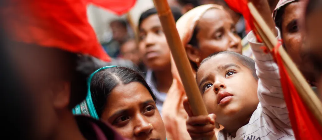 A Bangladeshi boy helps to raise a red flag at a demonstration. He is surrounded by the crowd at the protest, mostly women with headscarves. Credit: Rainbow Collective