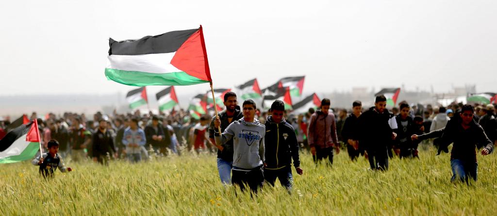 Palestinians take part in the Great March of Return in Gaza, Palestine, on Land Day, 30 March 2018. A crowd is gathered in a green field, holding Palestinian flags. Photo: Ashraf Amra/APA/Shutterstock