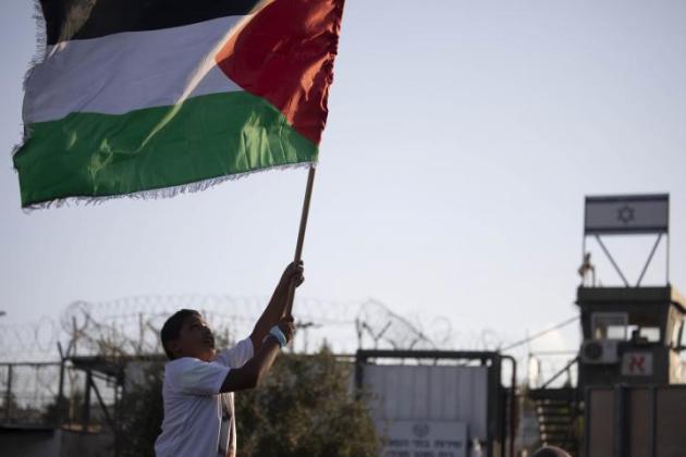 Boy runs with Palestinian flag in front of a prison with the Israel flag on.