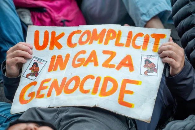 Person lying down holding a poster that reads, in bright orange text, 'UK COMPLICIT IN GAZA GENOCIDE'