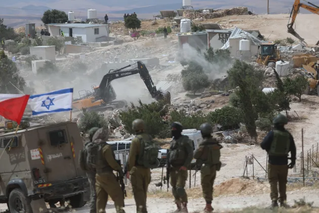 Israeli soldiers stand in the foreground while bulldozers and excavators demolish Palestinian structures in the village of Khallet Al-Dabaa in the West Bank. Dust and debris fill the air as heavy machinery destroys buildings, with several onlookers and structures visible in the background. An Israeli flag is mounted on a military vehicle nearby.