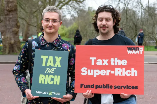 Two individuals stand side by side outdoors holding protest signs. The person on the left holds a green sign that reads "TAX THE RICH – EQUALITY TRUST." The person on the right holds a red sign that says "Tax the Super-Rich – Fund public services" with the logo "WAR ON WANT" in the corner. Trees and people in the background show the scene takes place in a park or public gathering area.