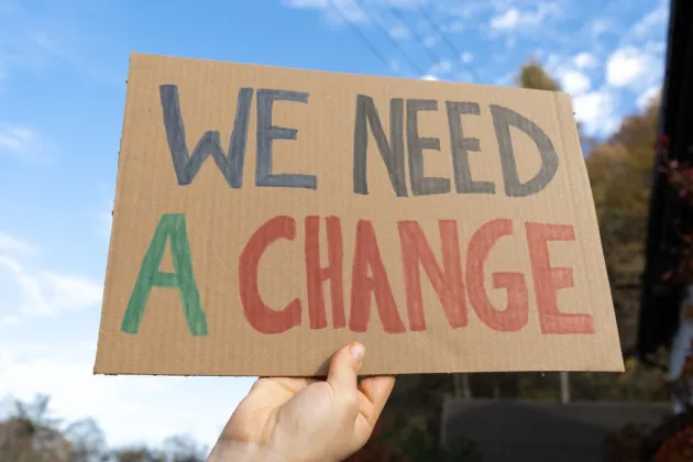 A person's hand holding a sign that says 'We need a change' in blue, red and green writing
