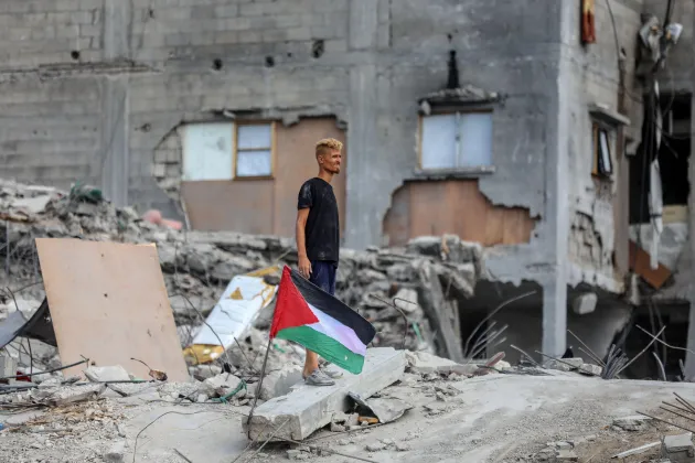 Man stands in rubble, next to a Palestinian flag