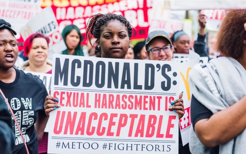 A Black woman at a demonstration faces the camera holding a sign that reads "McDonald's: sexual harassment is unacceptable #MeToo #FightFor15. She has black braids with a red streaks, tied up. Credit: FightFor15 Chicago