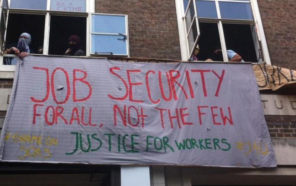 SOAS protesters with their faces covered hang a banner out of a window, reading "Job security for all, not the few. Justice for workers."
