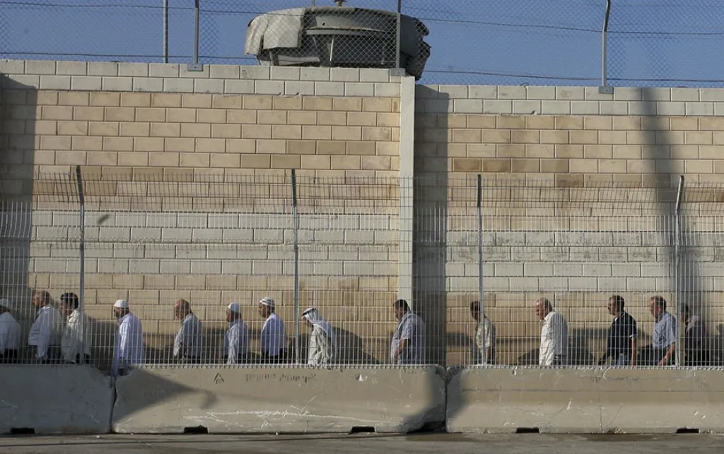 Palestinian workers in Israel protest dehumanising conditions at checkpoints. A queue of people stand in a line in front of a tall wall at a checkpoint. Source: Wikipedia