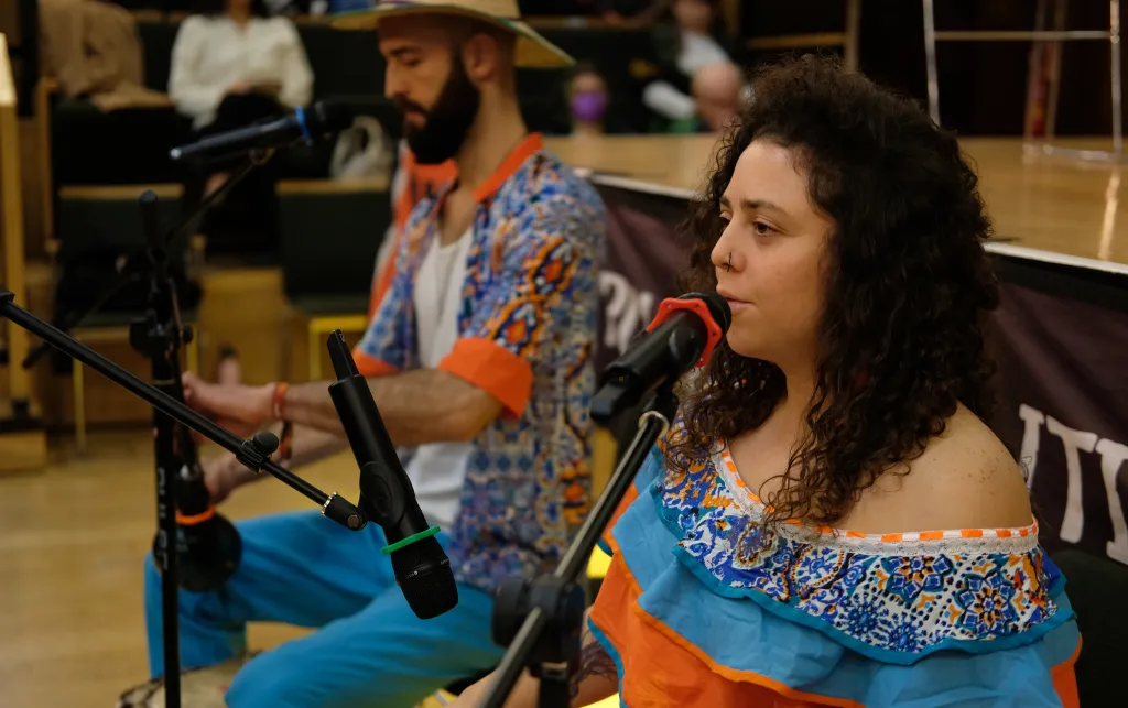 A close up of a man and a woman in the middle of a performance. Both have microphones in front of them which they appear to be singing into. The man is also holding a guitar.