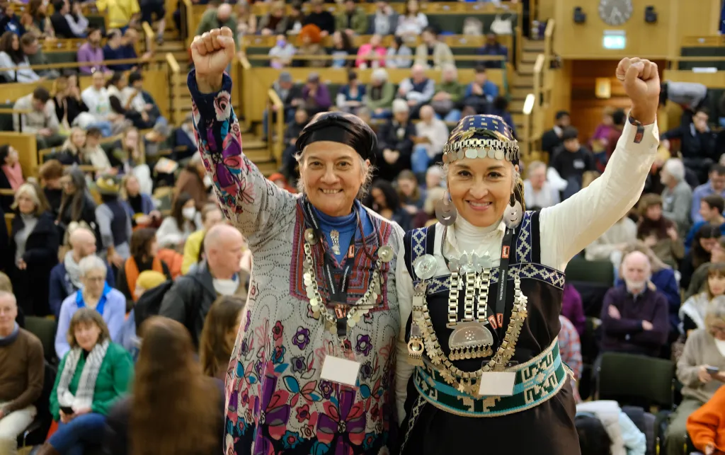 Photo of two women, Moira Millan and Vilma Diaz y Zarate. Both are smiling and have their fists raised in the air. Behind them we can see the audience in the auditorium.