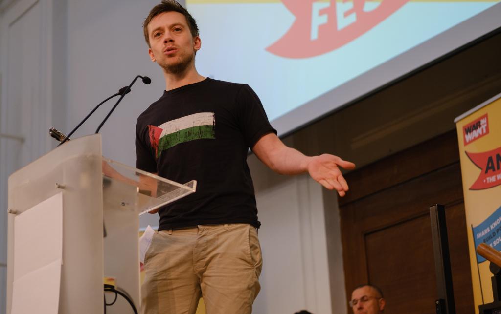 Owen Jones is pictured standing on a stage, in front of him is a lectern. He looks to be in the middle of a speech and has an animated expression on his face as he gesticulates.