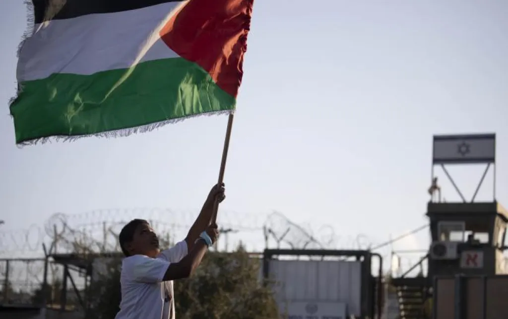 Boy runs with Palestinian flag in front of a prison with the Israel flag on.
