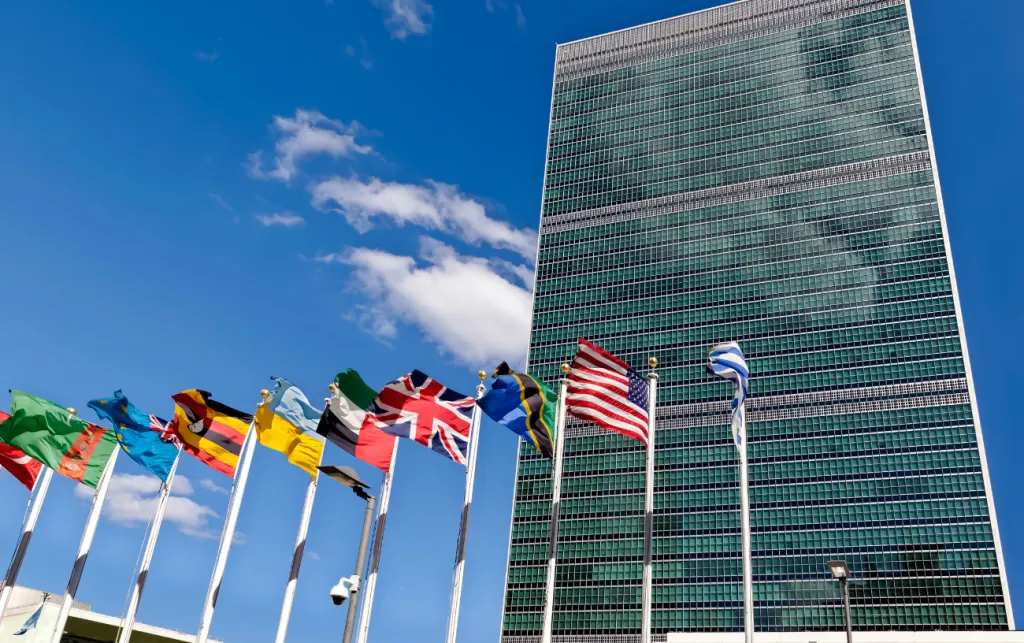 Tall glass building against blue sky with international flags waving in front.