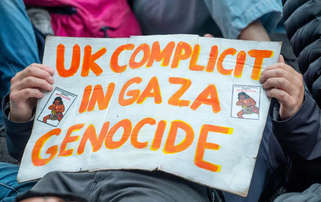 Person lying down holding a poster that reads, in bright orange text, 'UK COMPLICIT IN GAZA GENOCIDE'