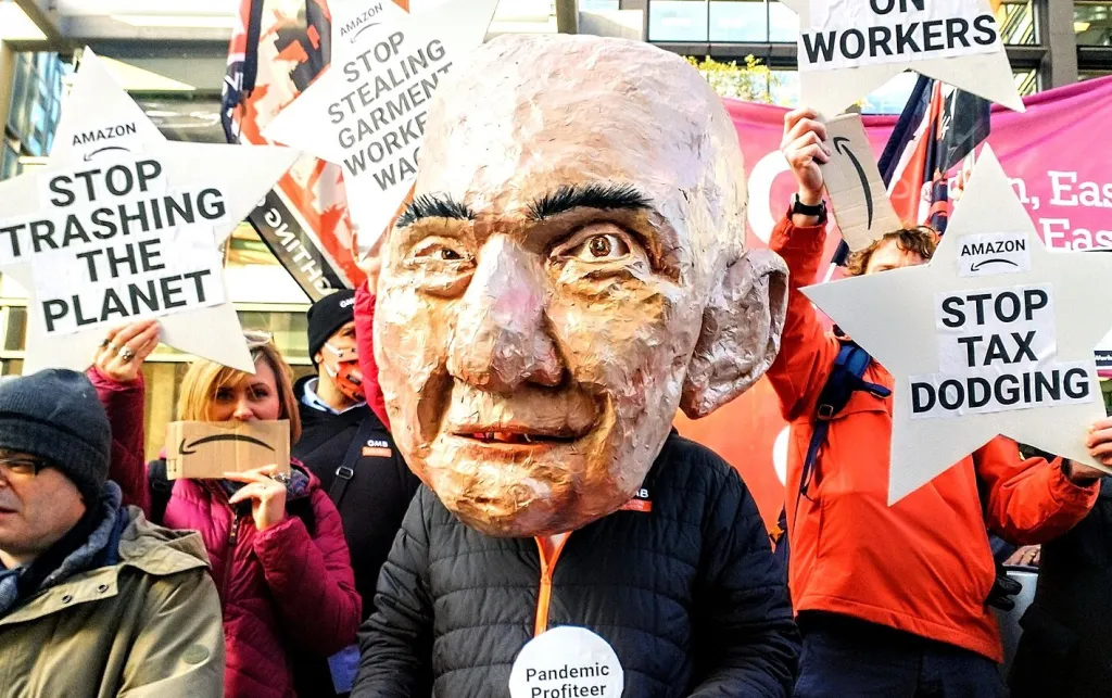 Paper mache head of Jeff Bezos worn by a protestor. Behind them many people hold banners saying phrases like 'Make Amazon Pay'.