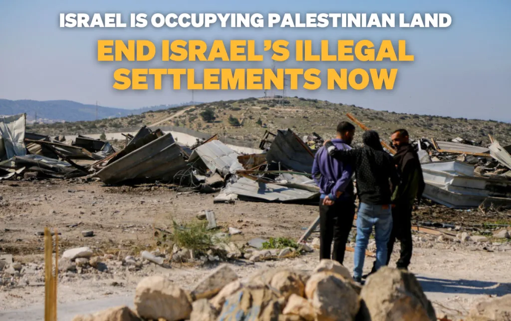 Three men embrace while looking at a destroyed building on arid land.