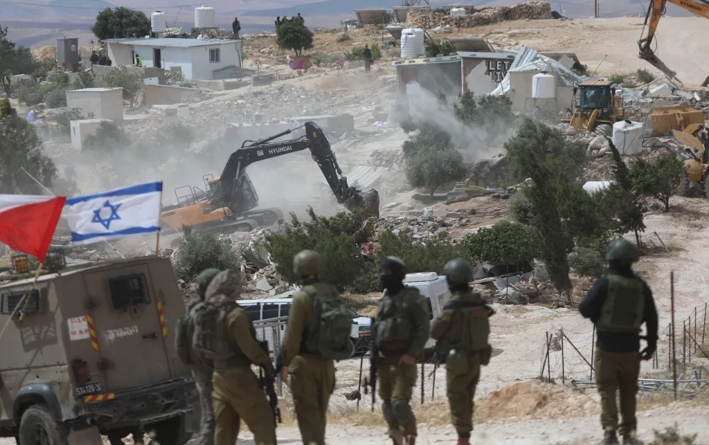 Israeli soldiers stand in the foreground while bulldozers and excavators demolish Palestinian structures in the village of Khallet Al-Dabaa in the West Bank. Dust and debris fill the air as heavy machinery destroys buildings, with several onlookers and structures visible in the background. An Israeli flag is mounted on a military vehicle nearby.