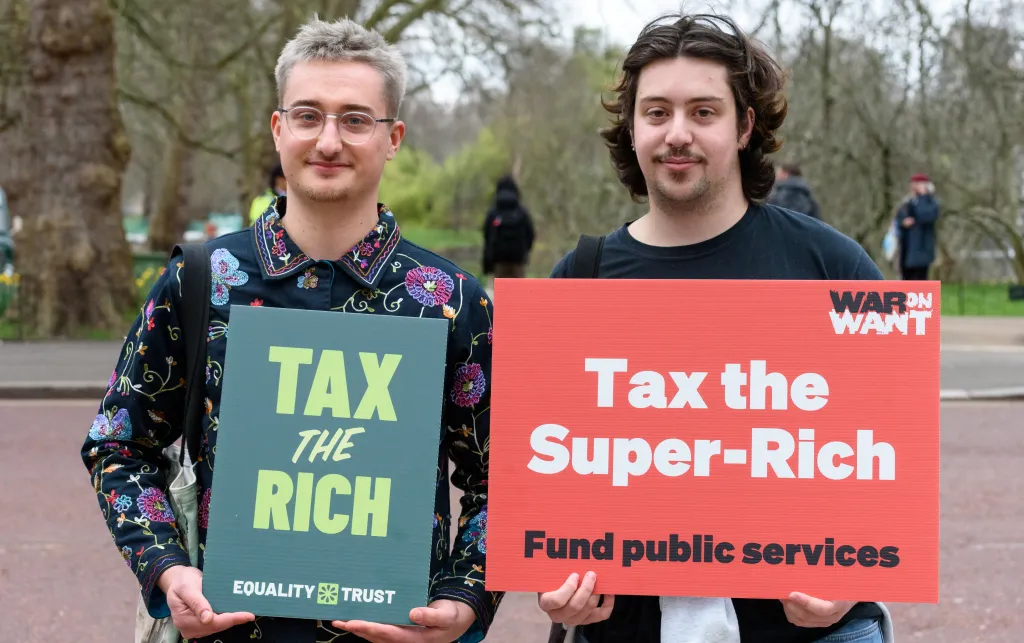 Two individuals stand side by side outdoors holding protest signs. The person on the left holds a green sign that reads "TAX THE RICH – EQUALITY TRUST." The person on the right holds a red sign that says "Tax the Super-Rich – Fund public services" with the logo "WAR ON WANT" in the corner. Trees and people in the background show the scene takes place in a park or public gathering area.