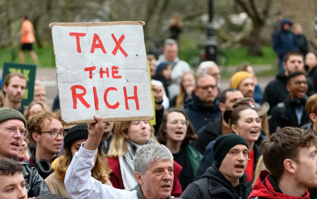 Man holds a banner saying 'tax the rich'. He is surrounded by people.