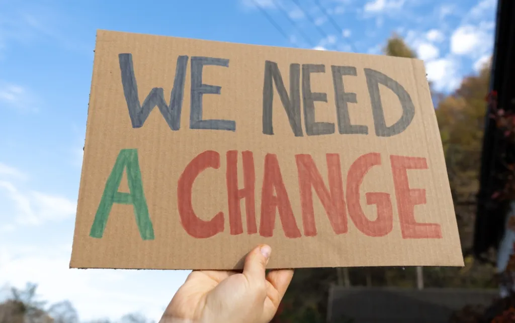 A person's hand holding a sign that says 'We need a change' in blue, red and green writing