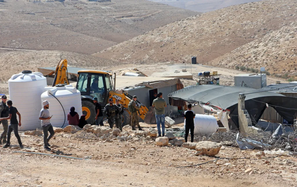 Yellow JCB bulldozer in small settlement with armed forces around