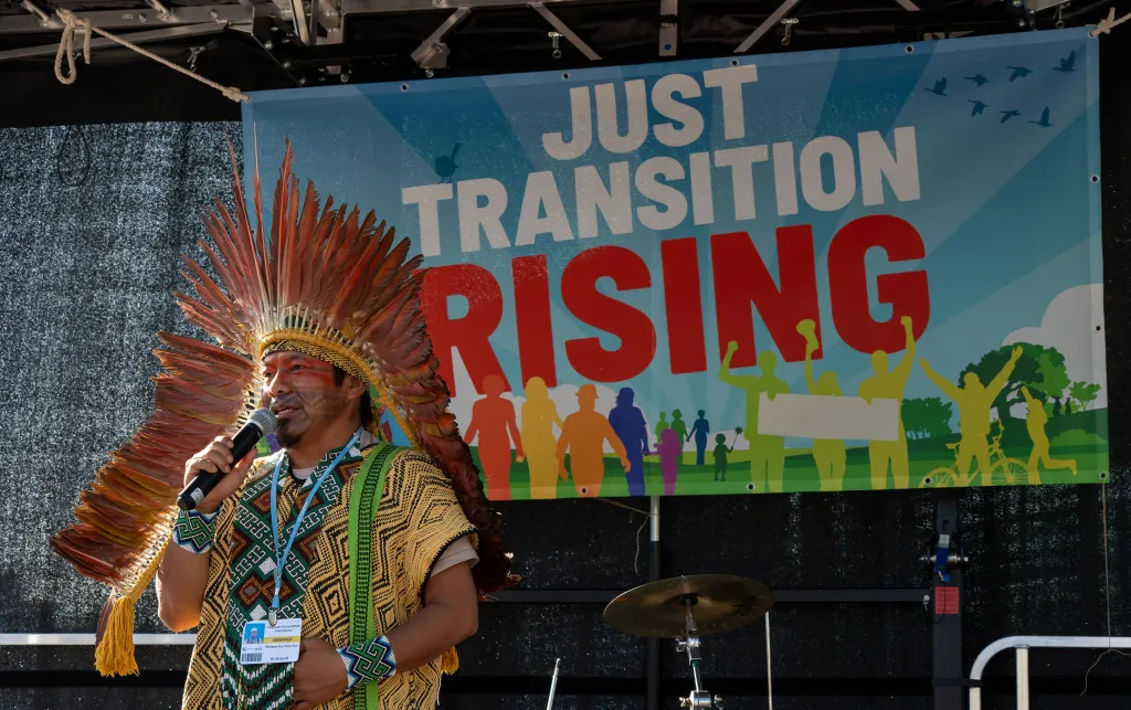 A man standing on a stage holding a microphone. Behind him is a colourful sign that reads: Just Transition Rising.