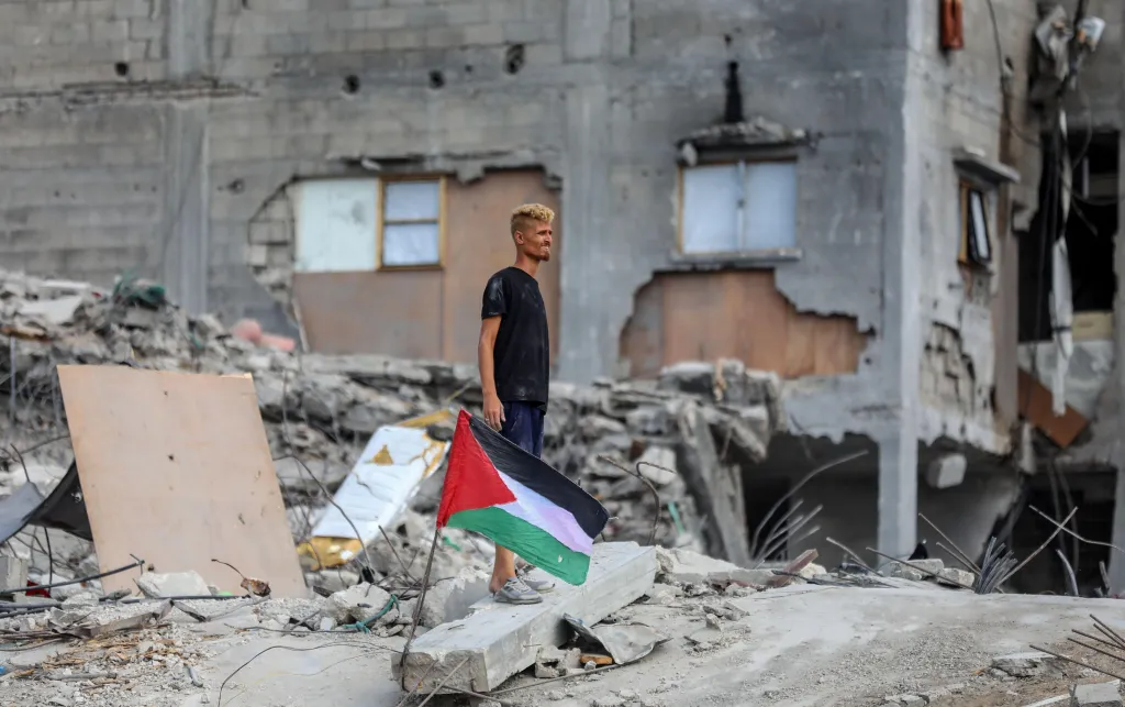 Man stands in rubble, next to a Palestinian flag