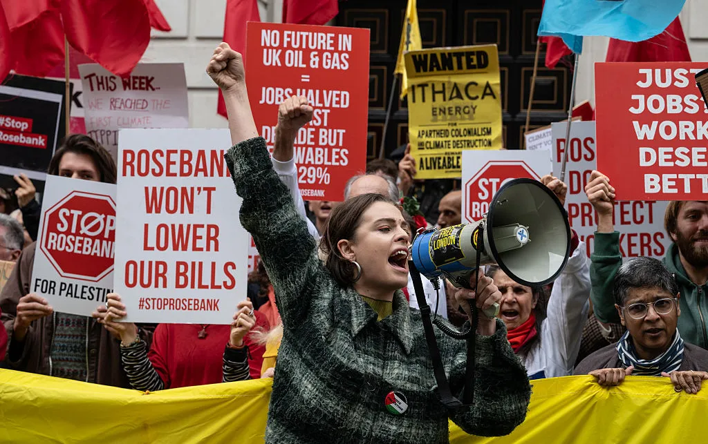 A woman is shouting into a megaphone at a protest. Behind her a lot of people are holding protest signs.