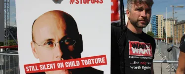 A War on Want campaigner at a demonstration holds a placard that has an image of G4S CEO Ashley Almanza's face with the words "Still silent on child torture" over his mouth, and #StopG4S in the top-right corner. Credit: War on Want