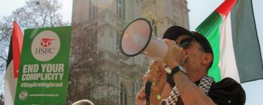 A protestor speaking into a megaphone in front of the Palestinian flag. A placard to the left reads "HSBC: End Your Complicity".