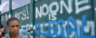 A black person standing in front of the fence at at a protest at Yarl's Wood Immigration Removal Centre. Graffiti on the fence reads: "No Borders! No one is illegal". Photo: IDJ Photography