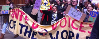 United Voices of the World (UVW) demonstration. A group of people, mostly women, march behind a UVW banner and hold placards, one of which reads "Reinstate Alba".