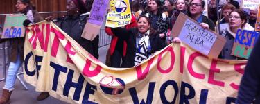 United Voices of the World (UVW) demonstration. A group of people, mostly women, march behind a UVW banner and hold placards, one of which reads "Reinstate Alba". 