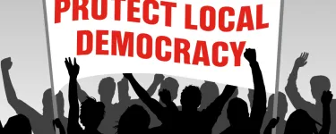 An illustration of a crowd in black and grey, holding up a white banner with red type that reads 'PROTECT LOCAL DEMOCRACY'.