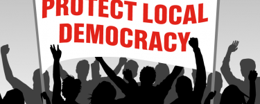 An illustration of a crowd in black and grey, holding up a white banner with red type that reads 'PROTECT LOCAL DEMOCRACY'.