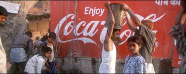 People standing in front of a Coca-Cola advert. One person with a white vest an moustache is passing a large drum that he is carrying on his head to another person.