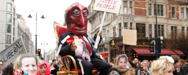 Protestors carrying a figure on a palanquin. The figure is wearing a devil's mask and carries a sign which reads 'Tax is for the little people'.