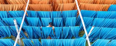 Man hangs up blue cloth on a line to dry. He is surrounded by similar blue and orange cloths. 
