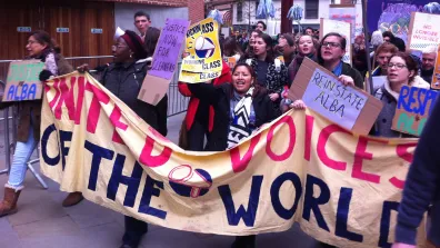 United Voices of the World (UVW) demonstration. A group of people, mostly women, march behind a UVW banner and hold placards, one of which reads "Reinstate Alba".