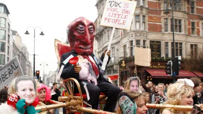 Protestors carrying a figure on a palanquin. The figure is wearing a devil's mask and carries a sign which reads 'Tax is for the little people'.