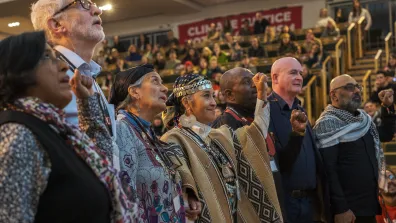 Photo of speakers standing in a row, looking upwards. Some have their fists raised in the air. Behind them we can see crowds of people sitting in an auditorium.