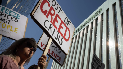 A person attending a protest. They are holding a protest sign which reads: "Cost of greed crisis".