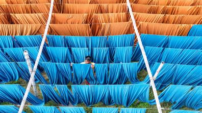 Man hangs up blue cloth on a line to dry. He is surrounded by similar blue and orange cloths. 