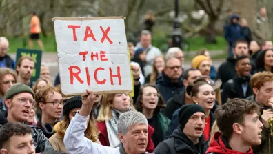 Man holds a banner saying 'tax the rich'. He is surrounded by people.